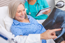 a woman patient lying in her hospital bed wearing a cheerful blue johnny as her doctor holds up an Xray while explaning what is shown. A nurse holding a clipboard stands at the other side of her bed.